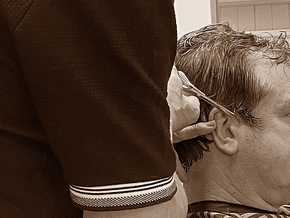 Barber giving a haircut to a middle-aged man in a classic barbershop setting with a patterned cape and wall clock in the background.